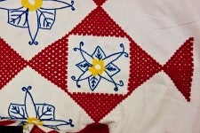 A white sheet with checkered red squares. In the white squares are blue and yellow embroidered flowers.