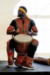 Alseny wearing bright orange, green and black colored traditional clothing sitting down while playing the drum. 